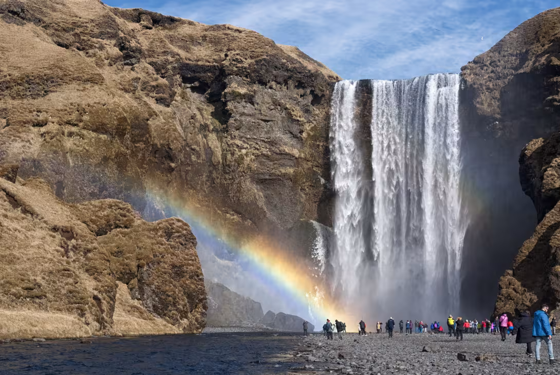 A group of people standing at the base of the waterfall with a rainbow over them.